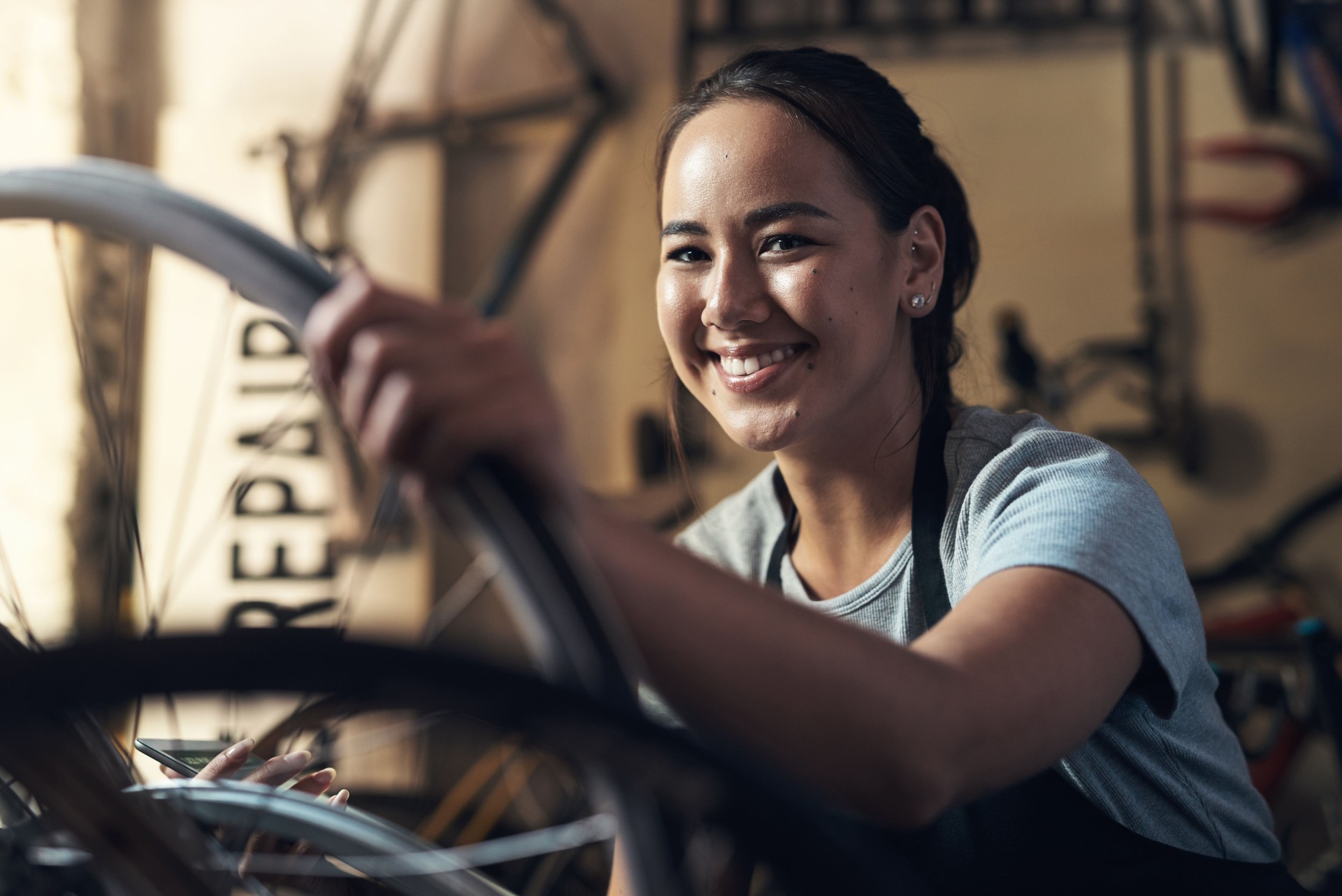 Retrato de una joven que se ve extremadamente complacida mientras sostiene un teléfono celular y arregla una bicicleta en un taller de reparación de bicicletas